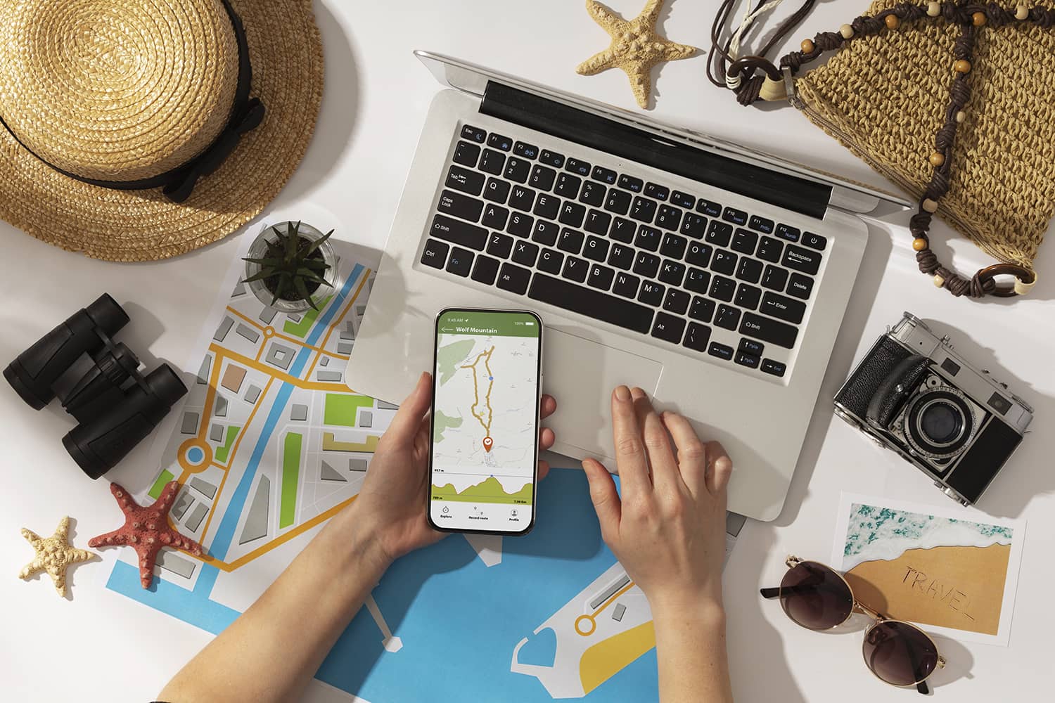 Persona planificando un viaje con un ordenador portátil y un smartphone mostrando un mapa, rodeada de objetos como un sombrero, prismáticos, estrellas de mar, una cámara, un bolso de paja, gafas de sol y una postal con temática de viaje.