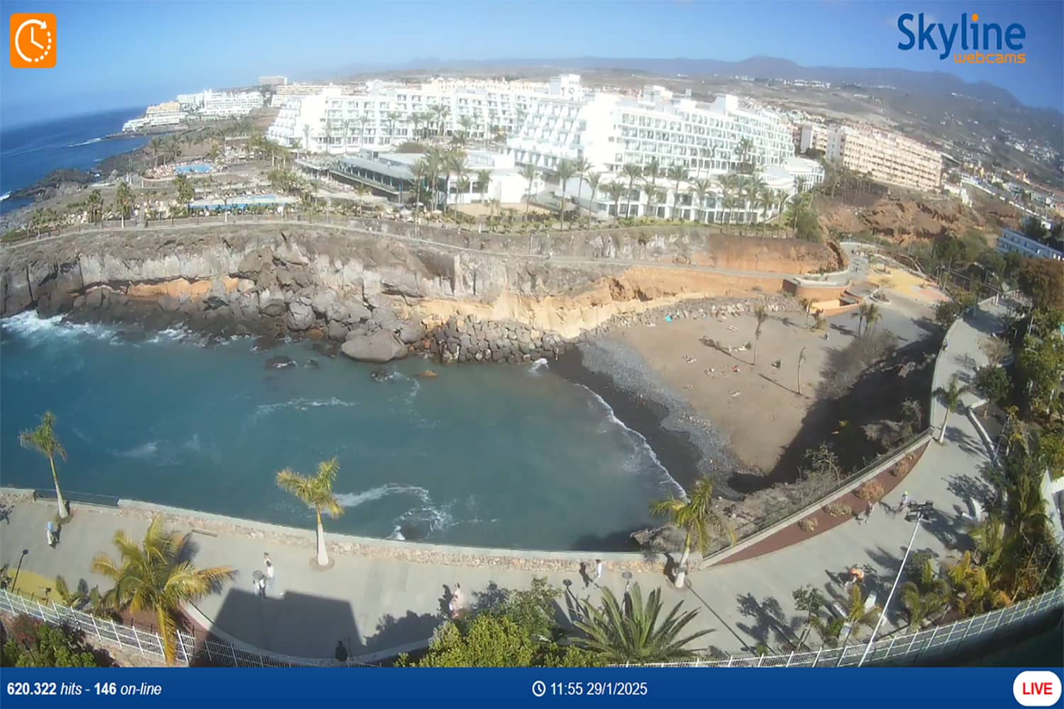 Una vista costera muestra una pequeña playa con poca gente, rodeada de acantilados rocosos. Al fondo, se ve un gran complejo hotelero blanco. Palmeras bordean un paseo marítimo con vistas a la playa. Se ve una interfaz de cámara con detalles de transmisión en vivo.
